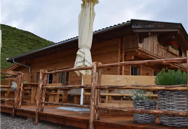 Eine gemütliche Holzhütte mit einer Terrasse aus Naturholz. Im Hintergrund sind grüne Hügel und ein bewölkter Himmel zu sehen.