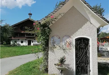 A small chapel with a cross and a beautiful garden. In the background, a traditional country house is visible.