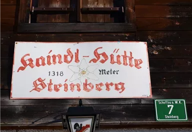 A sign with the inscription "Kaindl-Hütte" and the elevation "1318 meters" in Steinberg. In the background, a wooden building is visible.