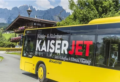 A yellow bus with the inscription "KaiserJET" drives through a picturesque landscape. In the background, mountains and a cozy alpine hut can be seen.