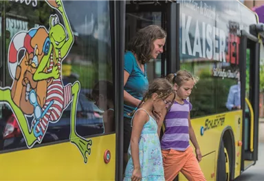 A woman and two girls are getting out of a colorful bus. The bus has a striking design featuring a drawing of a frog character.