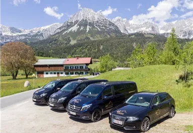 A group of vehicles is standing in front of a picturesque mountain panorama. In the background, green meadows and snow-covered peaks can be seen.