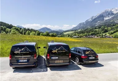 Three vehicles with the "Kaiser Taxi" logo are parked in front of a picturesque mountain landscape. In the background, green meadows and snow-capped mountains can be seen.