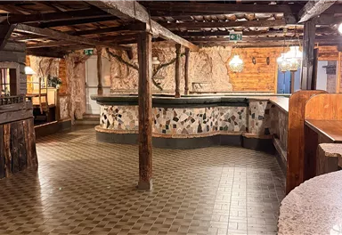 Ein rustikaler Raum mit Holzwänden und einem gefliesten Boden. In der Mitte steht ein Tisch und eine einfache Ausstattung, die eine gemütliche Atmosphäre schafft.
