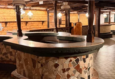 Ein rustikaler Raum mit einem großen, runden Steintisch in der Mitte. Die Wände sind aus Holz und die Böden sind gemustert.