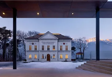 A historic building with a modern extension, surrounded by snow-covered trees. In the background, mountains and a clear sky are visible.