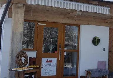 A cozy entrance to a wooden house with a sign saying "Kaiser Lidl". In front of the door is a table with pastries and a seating area.