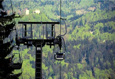 Eine Gondelbahn schwebt über grüne Berge und Wälder. Im Hintergrund sind sanfte Hügel und Bäume sichtbar.