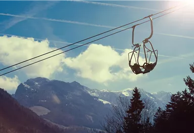 Eine Seilbahn schwebt über majestätische Berge unter einem klaren Himmel. Die Wolken und die Sonne verleihen der Szene eine malerische Atmosphäre.