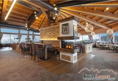 A modern lounge with wooden beams and large windows that offer a view of the mountains. Cozy seating and an open fireplace create an inviting atmosphere.