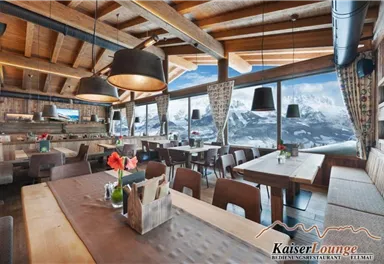 A cozy restaurant with rustic wooden interior and large windows that offer a view of the mountains. The tables are friendly decorated, and the atmosphere is inviting.