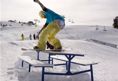 Ein Snowboarder macht einen Trick auf einer Tisch-Gelände im Schnee. Im Hintergrund sind Skilifte und weitere Fahrer zu sehen.