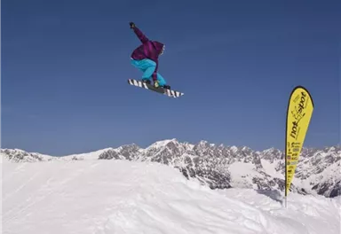 Ein Snowboarder springt in die Luft über einen schneebedeckten Hang. Im Hintergrund sind beeindruckende Berggipfel und ein gelbes Fahne zu sehen.