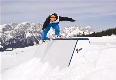 Ein Snowboarder springt über ein Hindernis in einer verschneiten Berglandschaft. Im Hintergrund sind mächtige Berge und ein blauer Himmel zu sehen.