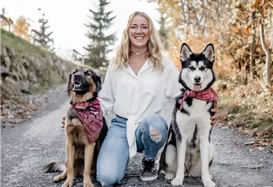 Eine Frau kniet auf einem Weg und posiert mit zwei Hunden. Der Hund links ist braun und der Hund rechts ist schwarz-weiß.