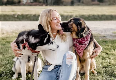 Eine Frau sitzt im Gras und umarmt zwei Hunde. Sie lächelt glücklich, während die Hunde liebevoll bei ihr sind.