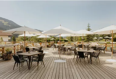 A cozy terrace with wooden tables and chairs. Large parasols provide shade and are surrounded by green landscapes.