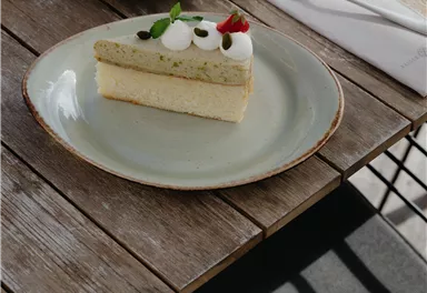 A piece of cake with a creamy layer and a green topping on a plate. In the background, a cup of coffee and cutlery are visible on a wooden table.