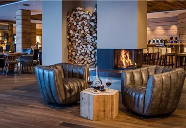 Cozy seating area with two leather chairs and a wooden table. In the background, a fireplace is burning and logs are stacked.