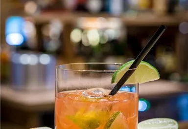 A refreshing cocktail in a glass, garnished with lime slices. In the background, a cozy bar atmosphere can be seen.