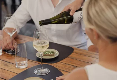 A couple is sitting at a table enjoying dinner. One person is pouring wine into a glass while small plates with food are on the table.