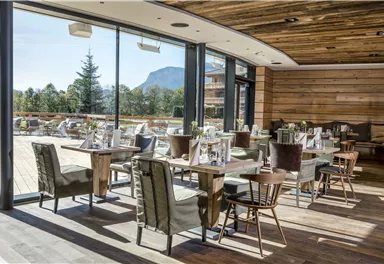 A modern restaurant with large windows and a view of nature. Cozy tables and chairs are arranged in an appealing setting.