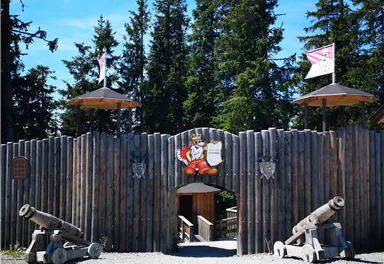 An entrance to a fortress with wooden stakes and two cannons. Colorful flags flutter above among tall fir trees.