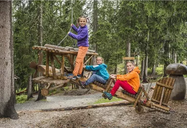 Three children are playing on a walkable wooden toy machine in the forest. They are wearing colorful jackets and laughing happily.