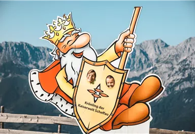 A cheerful cartoon figure of a king with a golden crown and a shield. In the background, impressive mountains can be seen.