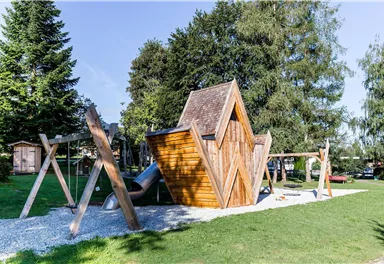 A playground with a modern wooden playhouse and a slide. In the background, tall trees and green grass can be seen.