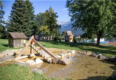 A playground with a water play area and a wooden bridge. In the background, trees and mountains can be seen.