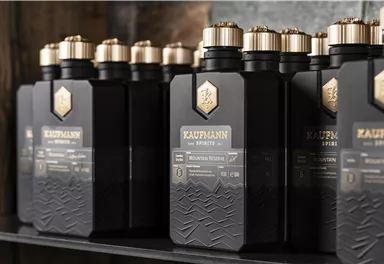 Eine Reihe von eleganten schwarzen Flaschen mit goldenen Verschlüssen steht auf einem Regal. Die Flaschen sind mit dem Namen "Kaufmann" und speziellen Produkten beschriftet.