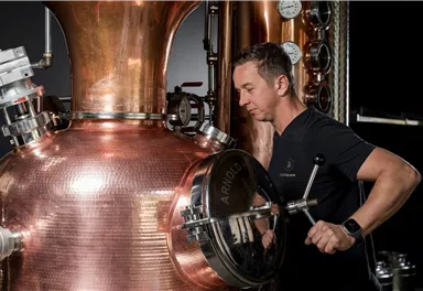 Ein Mann arbeitet an einer großen Kupfer-Destille. Im Hintergrund sind verschiedene Messgeräte sichtbar.