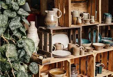 A stylish shelf wall made of wooden boxes with various pots and dishes. In the background, green plants are visible, creating a cozy atmosphere.