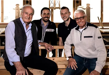 Vier Männer posieren lächelnd in einem Holzwerkstatt-Umfeld. Sie sitzen auf einer Holzbank und wirken freundlich und entspannt.