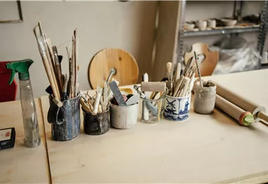 A workshop with various tools and brushes in containers. The table is made of light wood and in the background, other materials are visible.