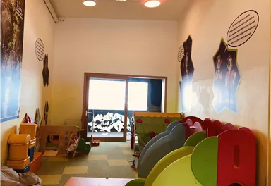 Ein heller Spielraum für Kinder mit bunten Spielgeräten. An den Wänden befinden sich informative Wandtexte und Fenster mit Blick nach draußen.