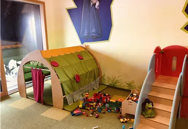 Ein Spielbereich für Kinder mit einem grünen Zelt, bunten Bausteinen und einer kleinen Rutsche. Der Raum ist hell und freundlich gestaltet.