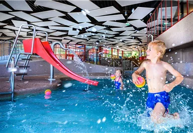 Ein Kinderbecken mit einer roten Rutsche. Zwei Kinder spielen im Wasser und spritzen fröhlich umher.