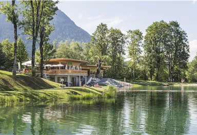 A picturesque building on the shore of a clear lake, surrounded by lush trees. In the background, gentle mountains can be seen, creating an idyllic atmosphere.