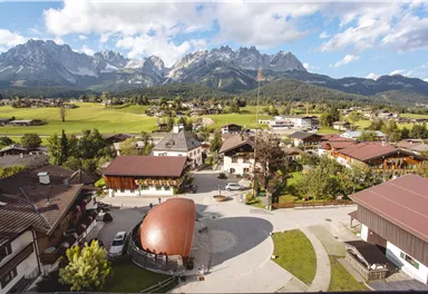 Eine malerische Landschaft mit einem kleinen Dorf im Vordergrund und beeindruckenden Bergen im Hintergrund. Der Himmel ist klar und die Natur ist grün und einladend.