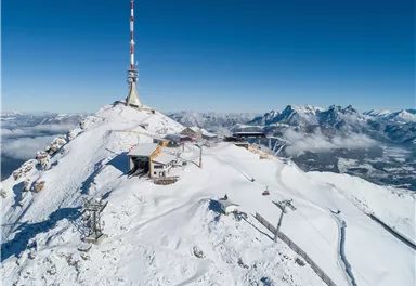 Ein schneebedeckter Berggipfel mit einem Sendemast und einer Berghütte. Im Hintergrund sind majestätische Berge und ein klarer blauer Himmel sichtbar.