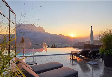 A beautiful pool area with loungers and a view of the mountains. The sun sets behind the mountains, bathing the scene in warm light.