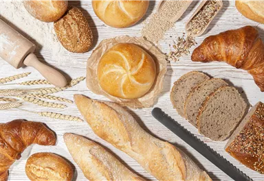 Eine Vielfalt von frischem Brot und Backwaren auf einer Holzoberfläche. Es sind Brötchen, Croissants und Scheiben von Vollkornbrot zu sehen.