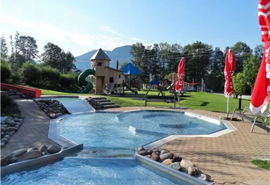 A beautiful pool area with a playground in the background. The surroundings are green and inviting, perfect for families.