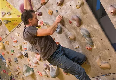 Ein Kletterer, der an einer Kletterwand mit verschiedenen Griffen unterwegs ist. Die Wand ist bunt und hat zahlreiche kleine Haltegriffe.