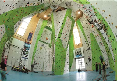 Eine Kletterhalle mit hohen Wänden und bunten Klettergriffen. Menschen klettern an den Wänden, während andere zuschauen und sich vorbereiten.