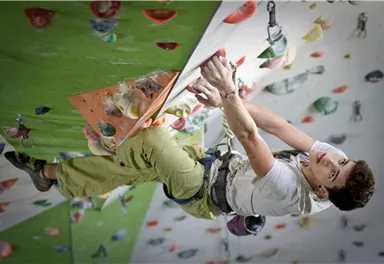 Ein junger Kletterer, der an einer Kletterwand mit bunten Griffen klettert. Der Sportler trägt einen Klettergurt und konzentriert sich auf den nächsten Griff.