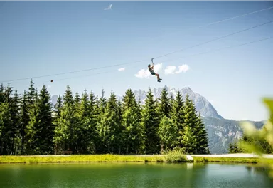 Ein Abenteurer fährt mit der Seilrutsche über einen klaren See. Im Hintergrund erhebt sich eine Berglandschaft mit dichten Wäldern.