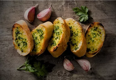 A plate with crispy garlic bread, garnished with fresh herbs. Next to the bread are some garlic cloves and parsley.